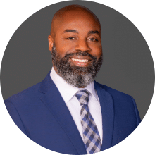 Professional headshot of Collin E in a suit and tie, smiling against a gray background.